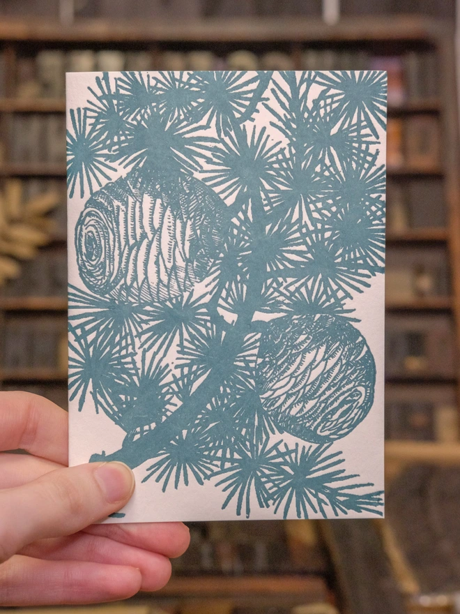 Card held up in situ. Design on front of card visible. Flat shaded artwork of cedar branch.