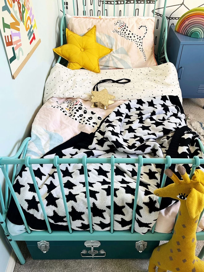 A view of a bright and colourful childrens bedroom in muted rainbow colours showing a turquoise metal framed single bed with a black and white star blanket draped and ruffled spread out on top of the duvet.