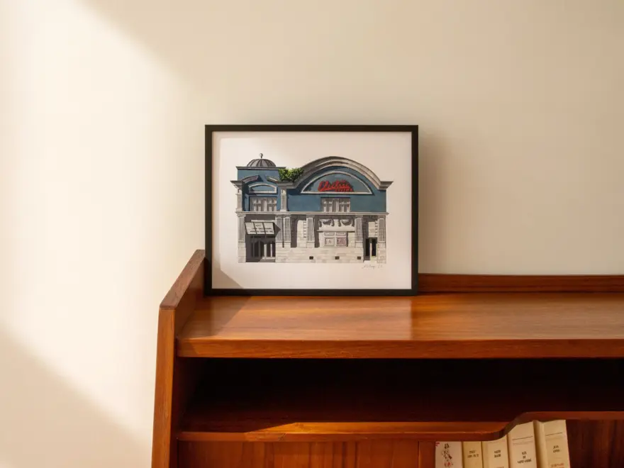 Giclee print of an illustration of the Electric Cinema in notting hill (a blue building on white background). In a black frame and sitting atop a dark wood desk, with white wall in background