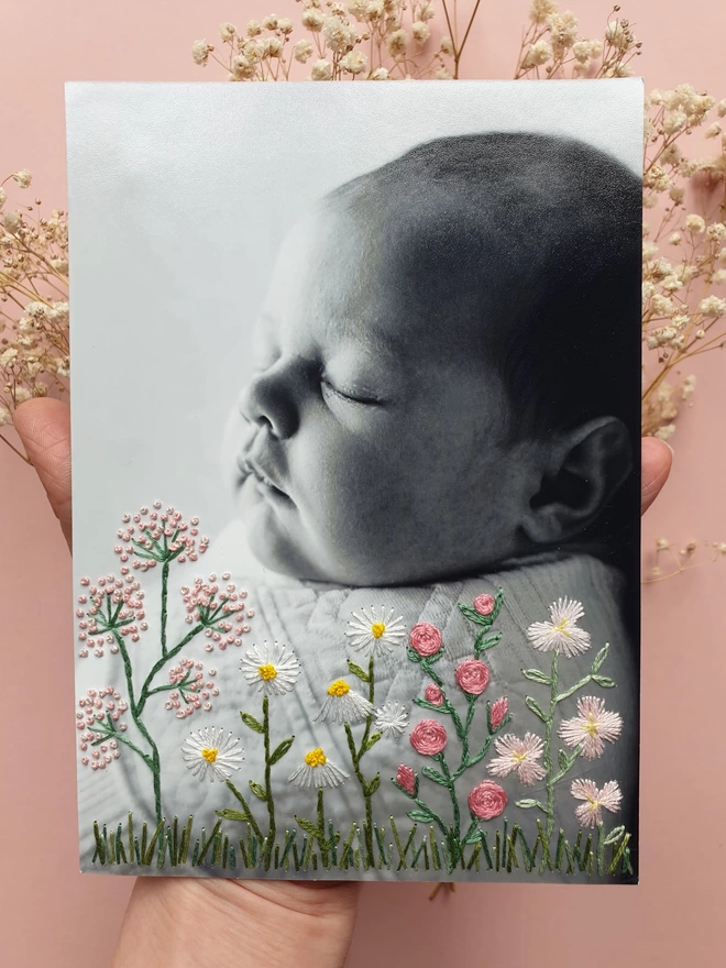 Baby photo with embroidered flowers growing from bottom of photo, held over pink background