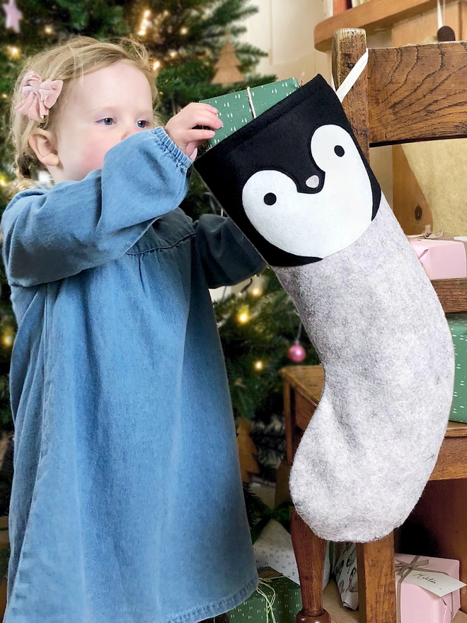 A young girl wearing a denim dress stands in front of a Christmas tree pulls a present out of a handmade felt penguin stocking.