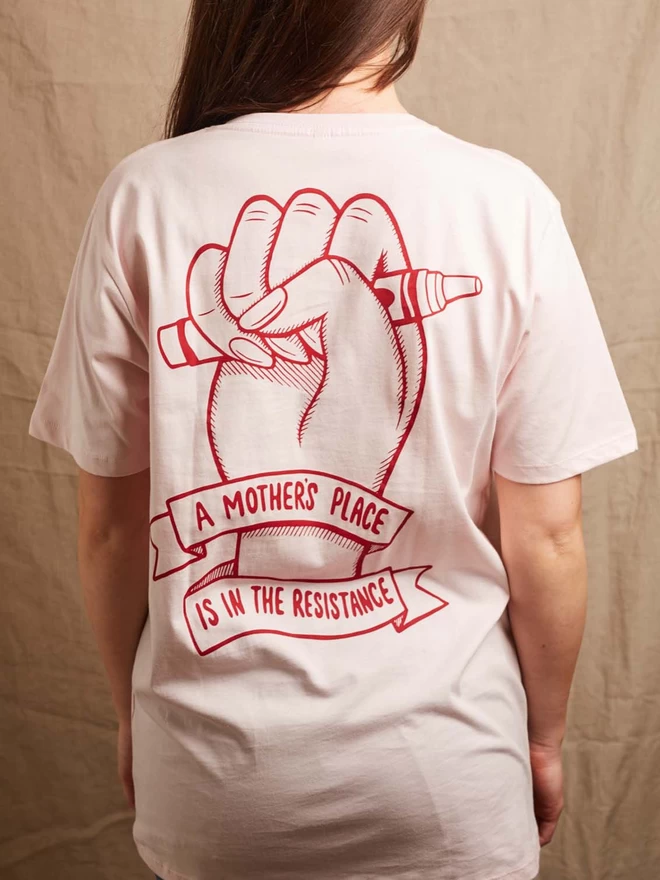 Model is showing the back of the Black & Beech pink A Mother’s Place is in the resistance t-shirt. The white print on the back shows a fist clutching a crayon and the with the words A Mother’s Place is in The Resistance written on a ribbon wrapped around the wrist 
