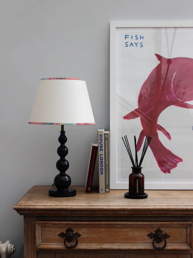 Empire lampshade with bold, colourful stripe trim, styled on a dark blue table lamp with books and artwork. A striped lampshade that blends classic empire light shades with a modern colourful twist.