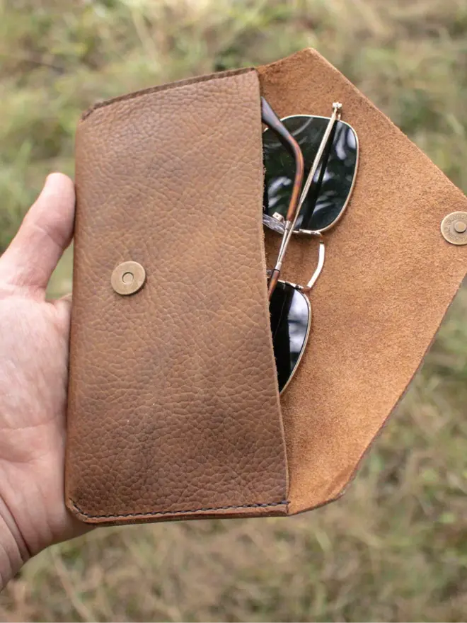 hand stitched leather pouch. a brown leather pouch held in hand against a grassy background, open to show sunglasses inside.