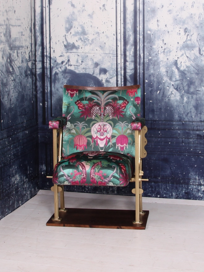 A single vintage cinema seat upholstered in a green and pink safari velvet, against a blue marbled wall.  The seat of the cinema seat is down