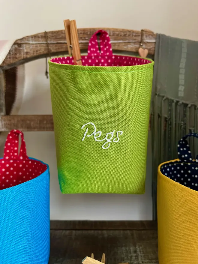 Green storage bag hanging on a wooden stand, the front embroidered with ‘Pegs’ in white thread