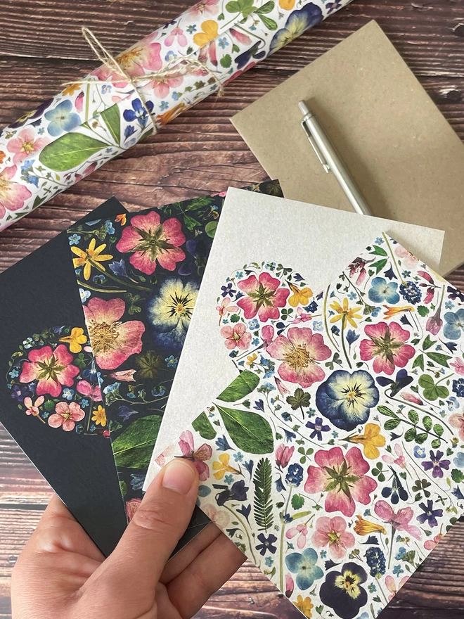 Hand Holding Set of 4 Floral Notecards with Pressed Flower Designs - Wooden Desktop with Envelope, Silver Parker Pen, and Pressed Flower Gift Wrap