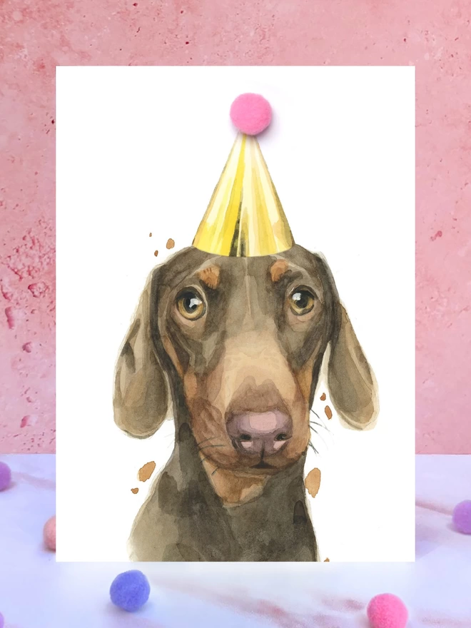 A greeting card featuring a hand painted design of a brown and tan dachshund, stood upright on a marble surface surrounded by pompoms. 