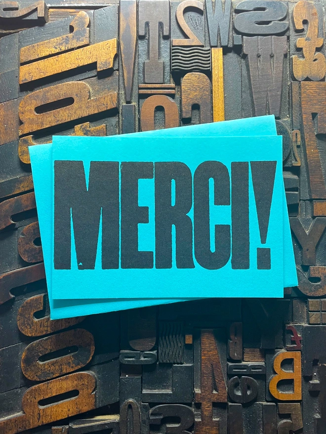 A bright turquoise thick duplex card with the word "MERCI!" printed in bold, black letters lies on a surface covered with various letterpress blocks.