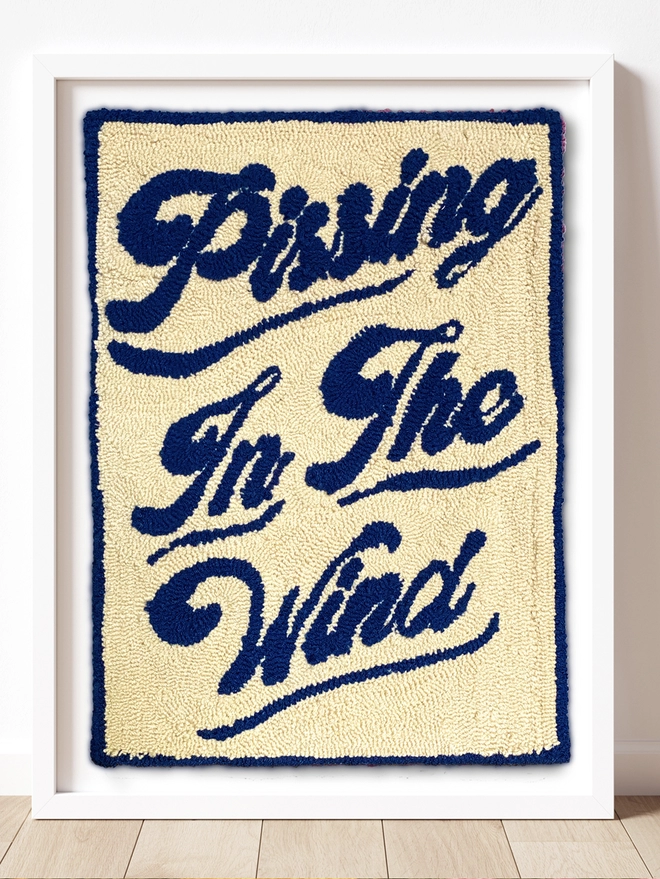 Pissing in the Wind written in navy blue wool on a cream wool backing.  Blue border around the image which is framed in a white box frame and sat on a wooden floor
