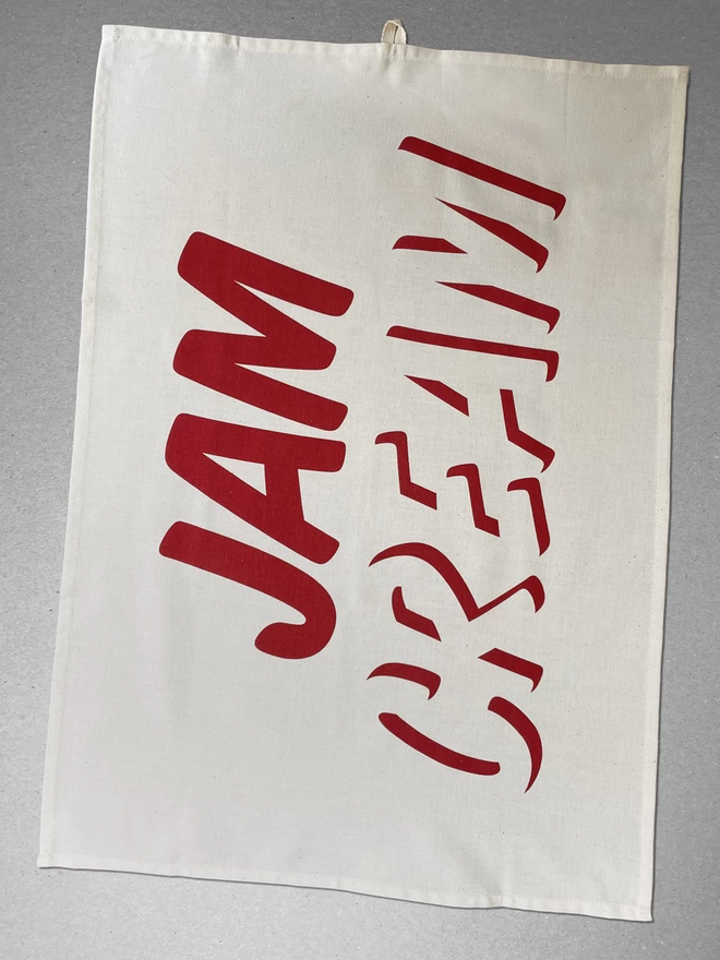 A flat view of the Cream First teat towel the Jam is in bold and the Cream is depicted by shadow around the letters, making it even ore creamlike. laid on a plain grey background.