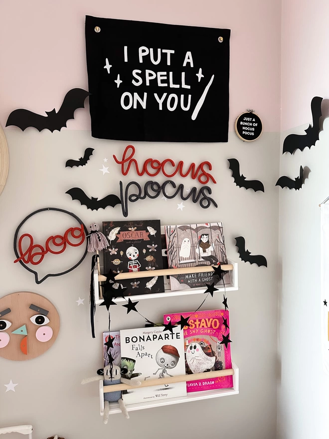 'Hocus Pocus' knitted wire words hanging up on the wall above a bookshelf full of Kid's Halloween Books with lots of Hallow's Eve decor surrounding it. 