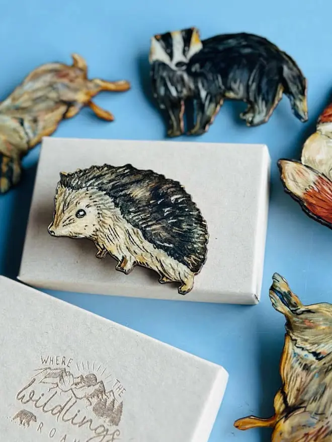 Hedgehog Brooch Wooden Accessory, placed on top of a white box, resting on a blue table. 