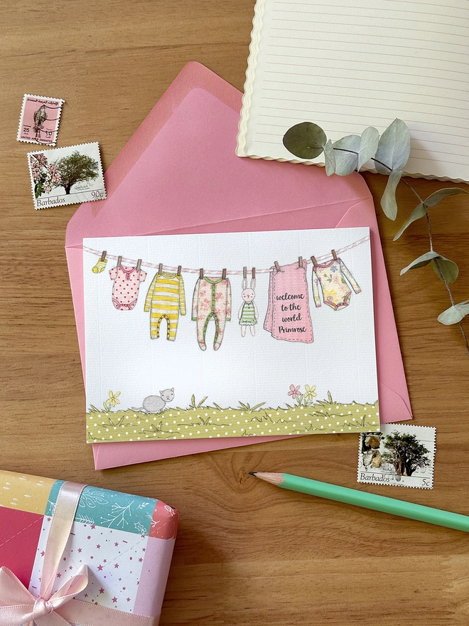 A personalised new baby card with an illustrated washing line of baby clothes lays on a pink envelope on a wooden desk.