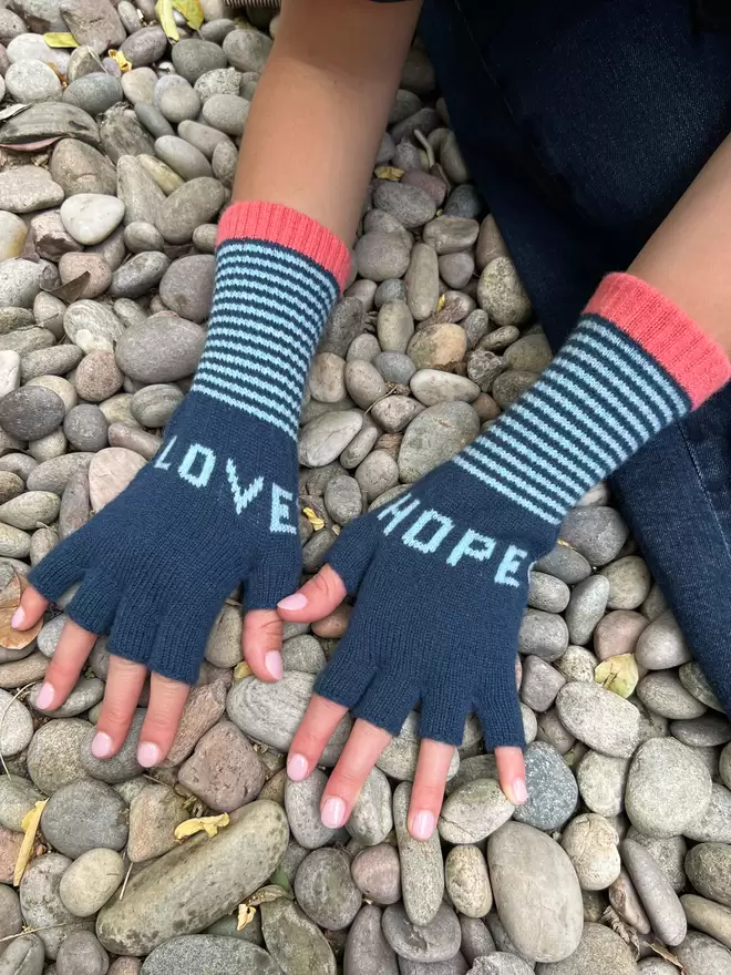 A teal fingerless knitted glove with the words LOVE and HOPE in pale blue and a stripe cuff with coral edge detail. Made from 100% Lambswool with Wool spun in Yorkshire. 