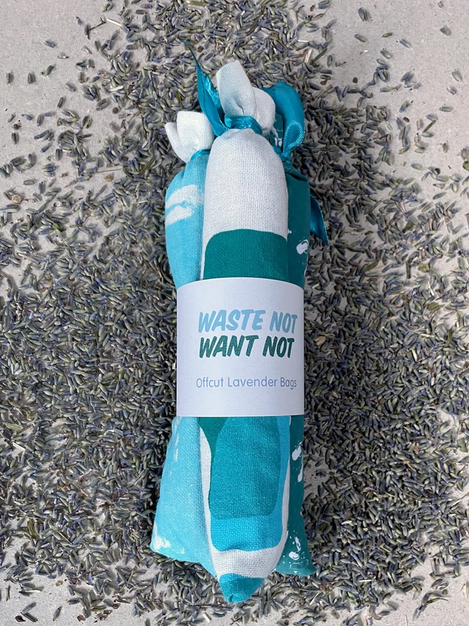 A bundle of three long style cotton lavender bags, tied at the tops, gathered with a branded cuff, sit on a scattered pile of fresh lavender - it says Waste Not Want Not, Offcut Lavender Bags. The fabric has bits of turquoise and teal pattern visible.