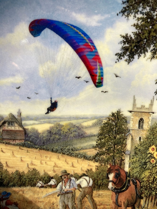 Close up of a paraglider sailing into a field as part of a rural scene on a vintage plate