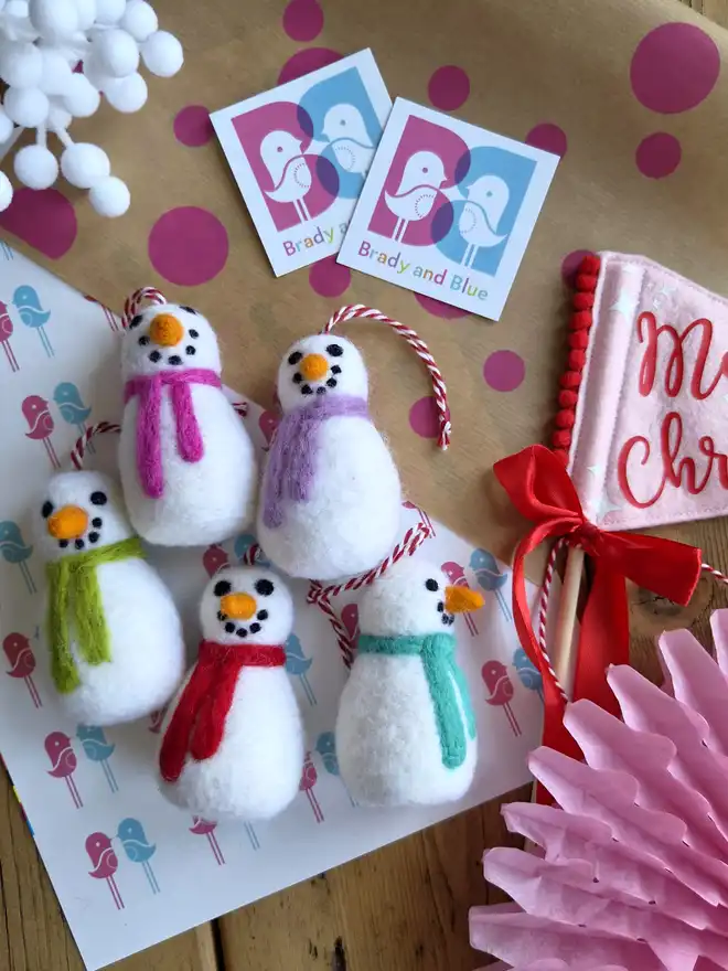 Felt snowmen tree decorations, with different coloured scarves - pink, turquoise, lilac, lime and red. Laid on a table with gift wrap and stickers.