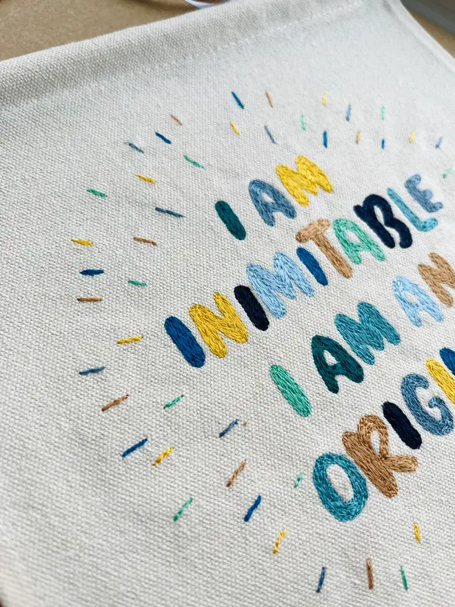 custom embroidered banner. cotton banner embroidered with 'i am inimitable i am an original' in colourful stitching.
