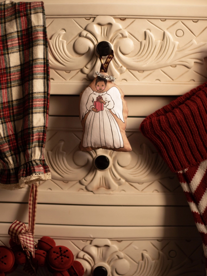 a Christmas tree angel depicting an asian small girl holding a red present made from fabric with a tinsel halo and ribbon for hanging hung on a white dresser between Christmas stockings