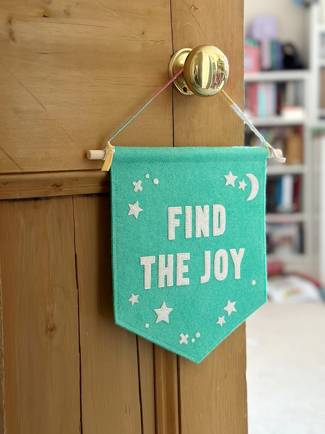 A turquoise felt banner with white words that read "Find The Joy" stitched on hangs from a ribbon hanger on a wooden door.