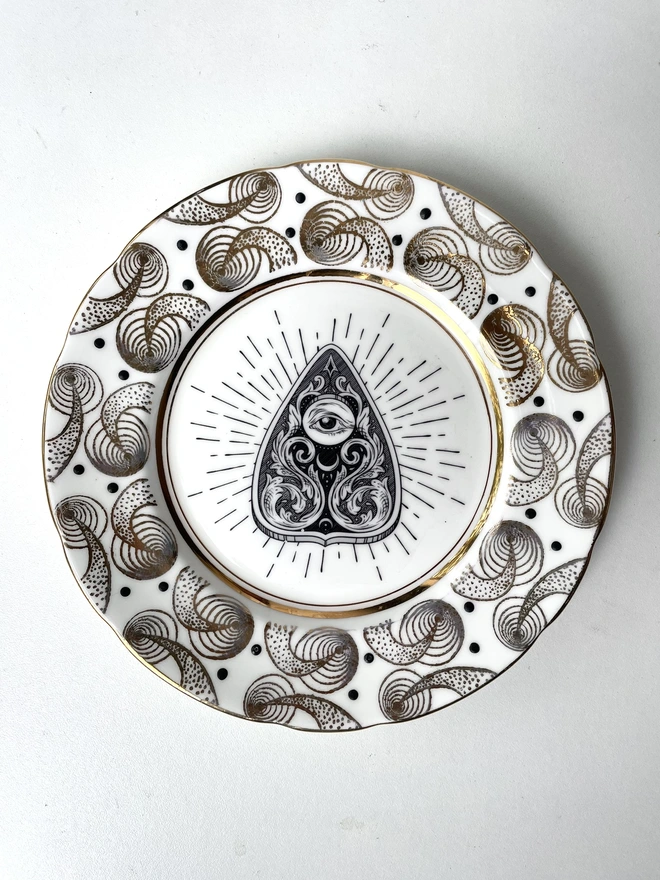 vintage plate with an ornate border, with a printed vintage illustration of a Ouija Planchette in the middle 
