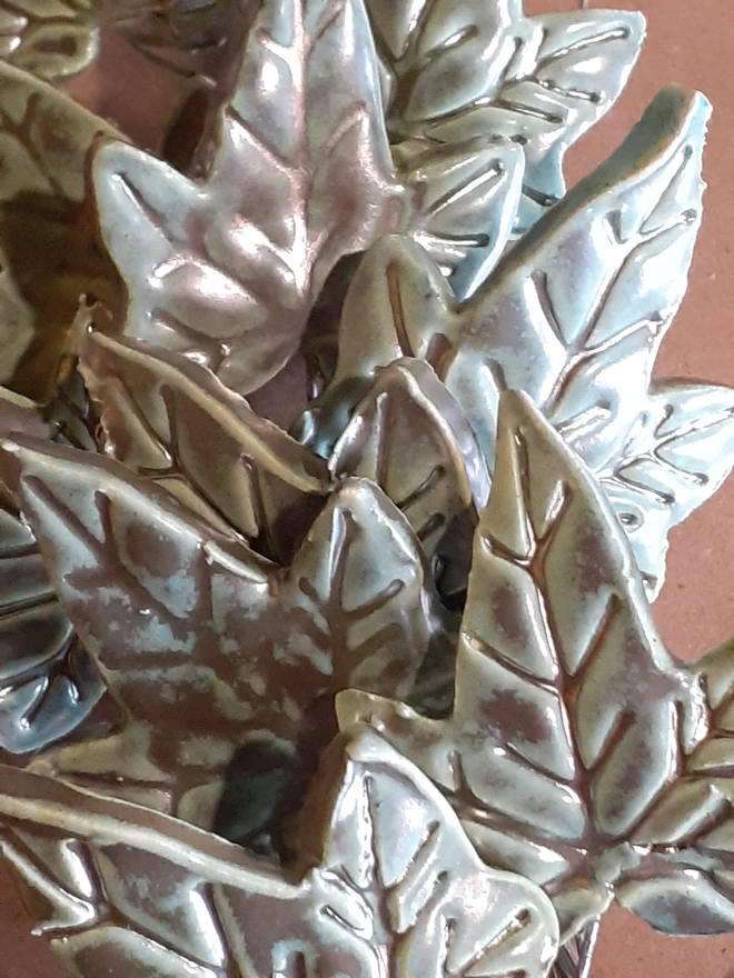 Close-up of overlapping handmade ceramic ivy leaves, showing their deeply impressed veins and glossy verdigris glaze with shimmering metallic copper highlights.