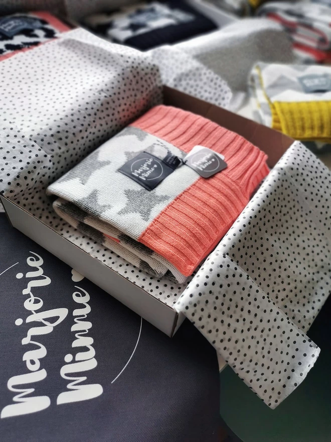 A neatly folded star blanket is shown packed inside a box and wrapped with tissue paper. The box is sitting on a table surrounded by other gift wrapped products. Beneath the box is a grey Marjorie Minnie branded gift bag.
