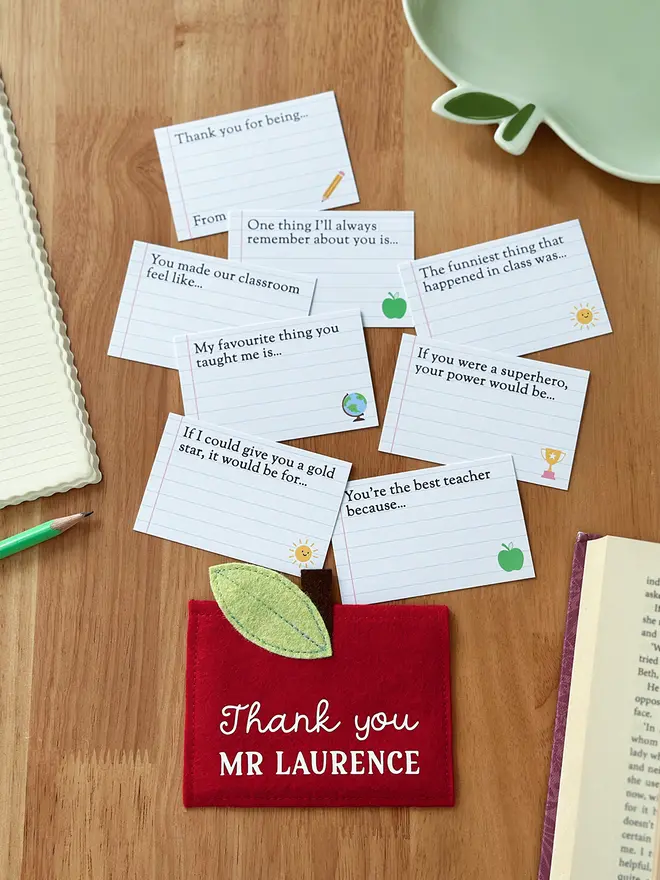 A handmade red felt apple-shaped envelope with "Thank you Miss March" written on the front. The heirloom apple envelope surrounded by completed prompt cards with handwritten messages from a child, displayed on a wooden desk. It's a unique personalised gift for teachers that is perfect for an end of term thank you.