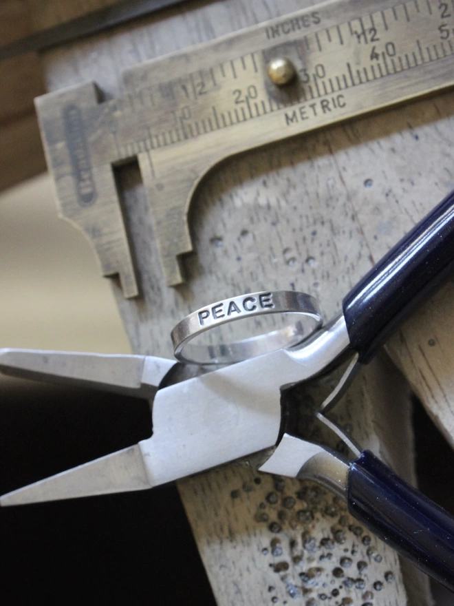 A sterling silver ring band with the word 'peace' stamped on it, resting against a pair of blue handled pliers on a jewellery bench, with a brass pair of callipers in the background.