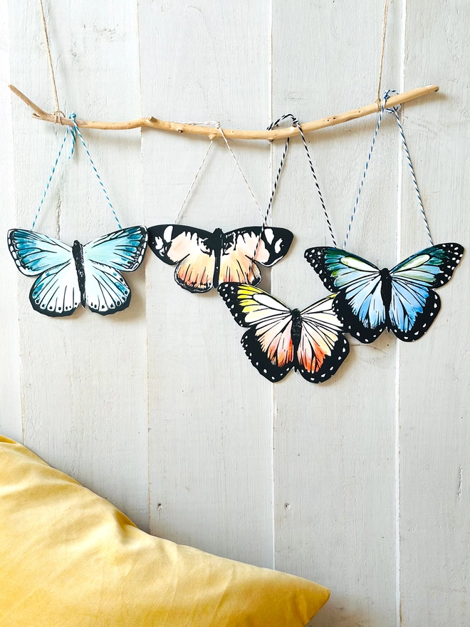 Hand Painted Butterfly Colourful Linocut Hanging Decoration In multiple colours and black four hang nect to each other on a stick for display with a yellow velvet cushion below