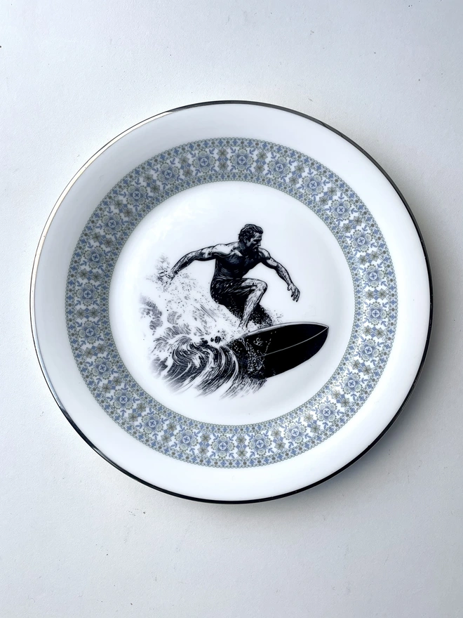 a vintage style image of a surfer riding a wave, 60s style, is printed in the centre of a vintage plate with an ornate edge
