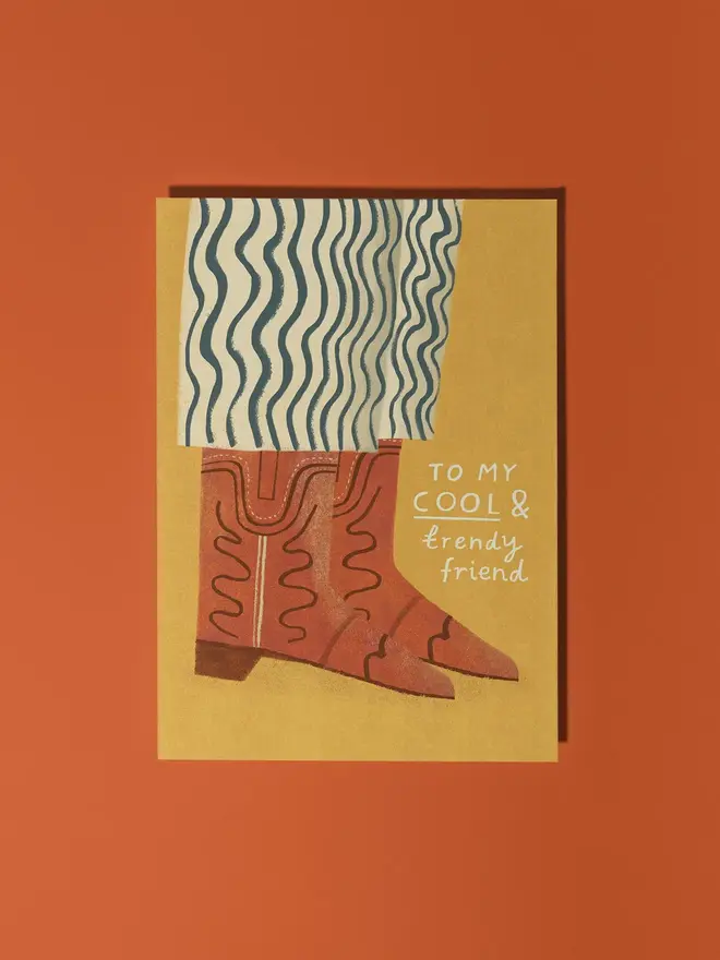 cowboy boots illustrated friendship card. brown cowboy boots worn with black and white striped trousers illustrated on a mustard yellow background, photographed against a brick red backdrop.