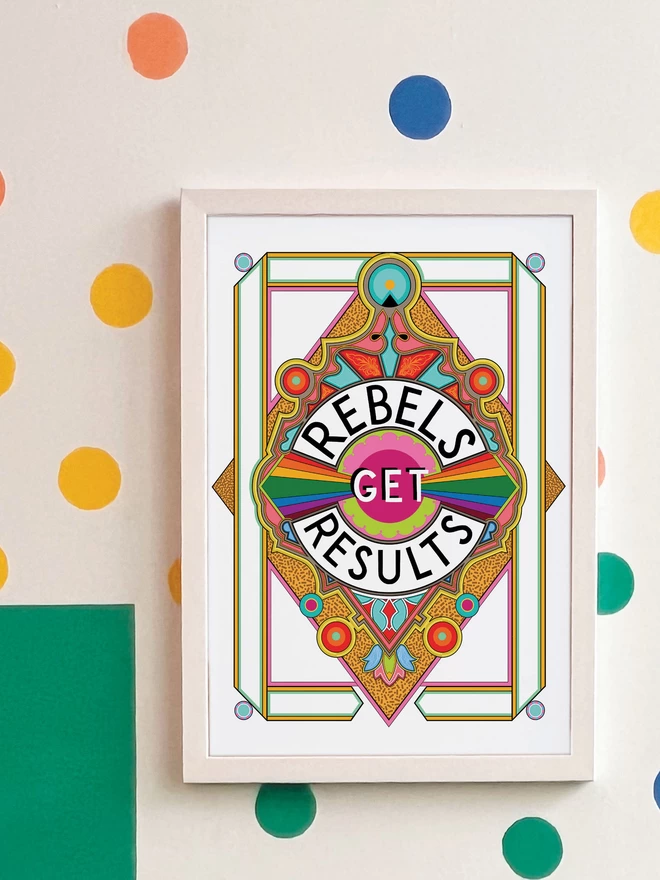 Rebels Get Results is written in black on a white background at the centre of this vibrant, abstract portrait illustration, with a white background and rainbows emitting from the centre, and multi-coloured detailing. The picture is hanging in a white frame on a white wall, with yellow, orange, green and blue spots and a green and yellow rectangle painted in the top left hand corner.