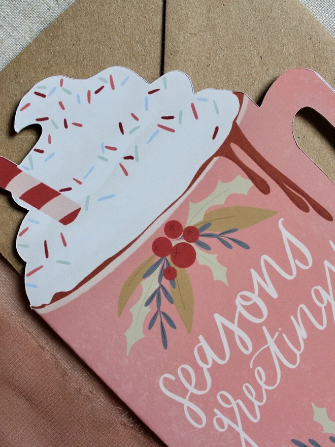 season's greetings christmas hot chocolate card. christmas card shaped like a pink mug with whipped cream on top.