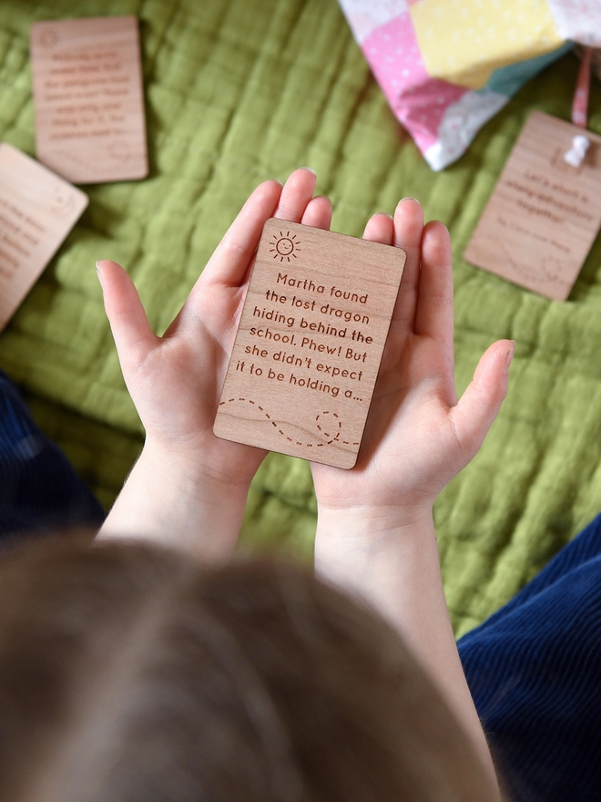 A young child holds a wooden story card with engraved wording on the palms of her hands above a green blanket. 
