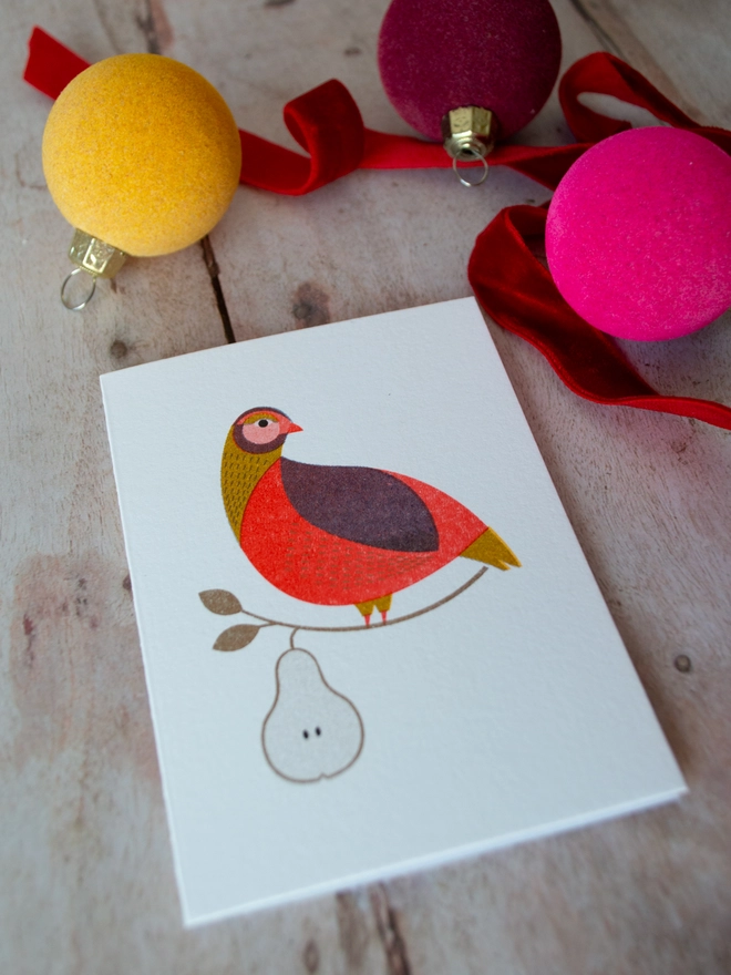 Bright and colourful, a risograph printed mini Christmas card featuring the traditional folklore character, a Partridge in a Pear Tree is placed on a wooden table with jewel toned festive Christmas baubles and ribbons surrounding it.