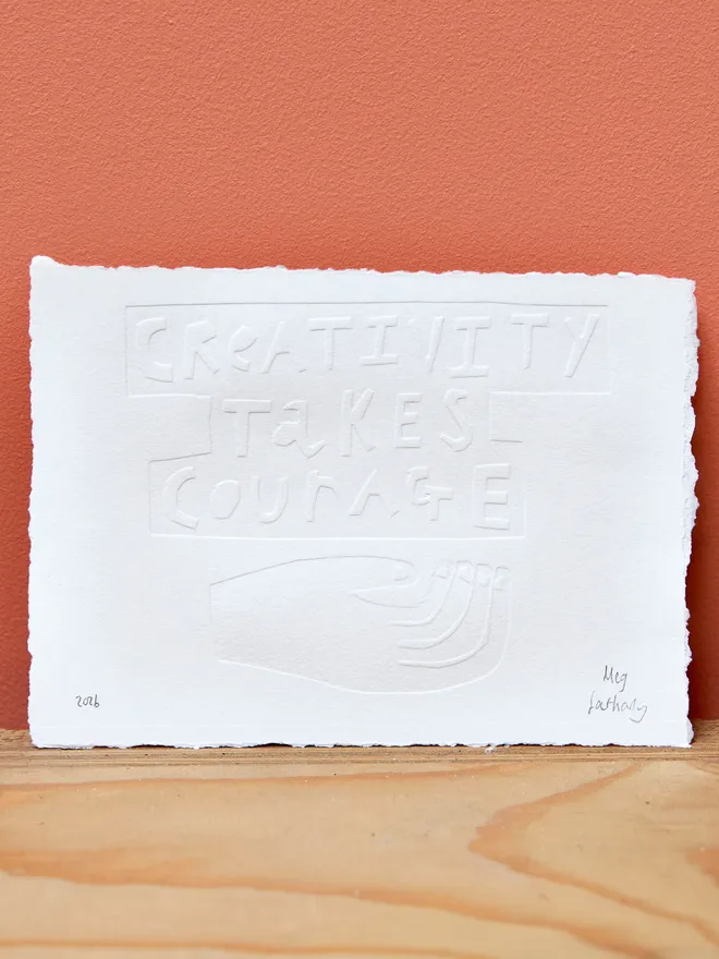 'Creativity Takes Courage' embossing on white paper, signed by the artist and held up against a coral wall atop a wooden surface