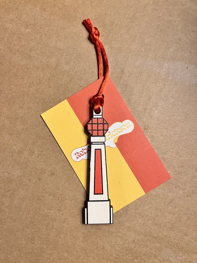 Small white and orange wooden tree decoration in the shape of Margate Lido sign. It is on a brown card background with a red ribbon and a small yellow and orange card saying 'Rebecca Strickson"