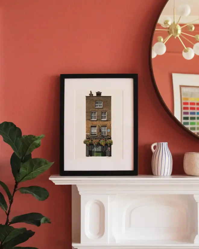 Limehouse The Grapes Giclée Print in a black frame, sitting on a white fireplace in front of a coral coloured wall, with plant to the left and a circular mirror and white vase to the right