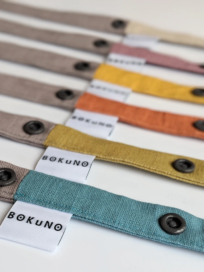 Handmade toy straps in soft neutral and pastel tones, featuring sturdy snap fastenings and small fabric tags with a bunny logo, neatly arranged in a row.
