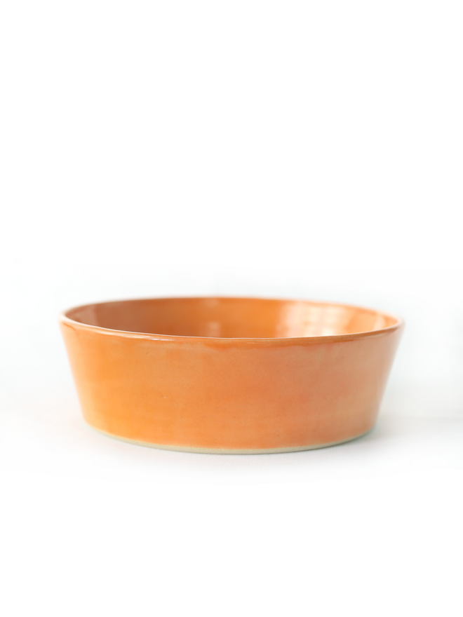 A single wide, shallow glazed ceramic pasta bowl in mandarin orange, photographed from a slight angle against a plain light grey background, showing the unglazed natural clay base.