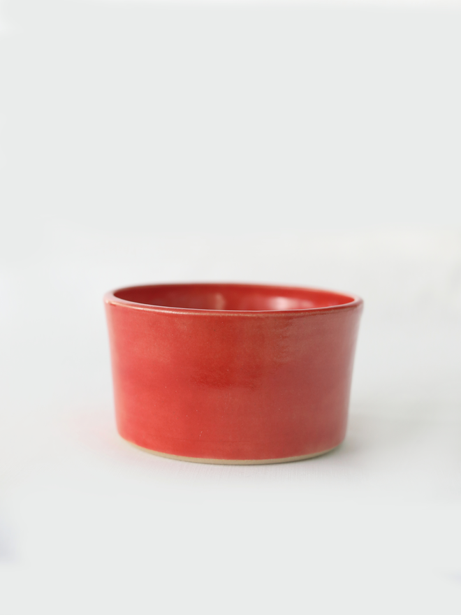 A small handmade ceramic side bowl in glossy cherry red glaze, with a tapered sides and a wide open rim, photographed against a plain light grey background