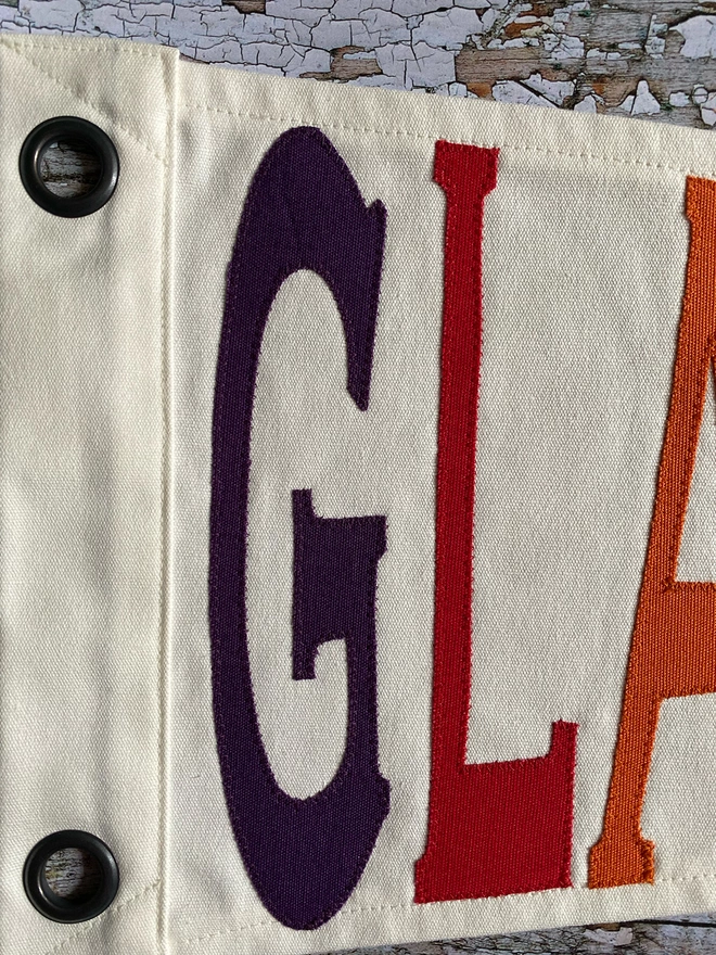 Detail of an ivory Glasonbury pennant flag showing the letters GLA in purple, red, and orange canvas