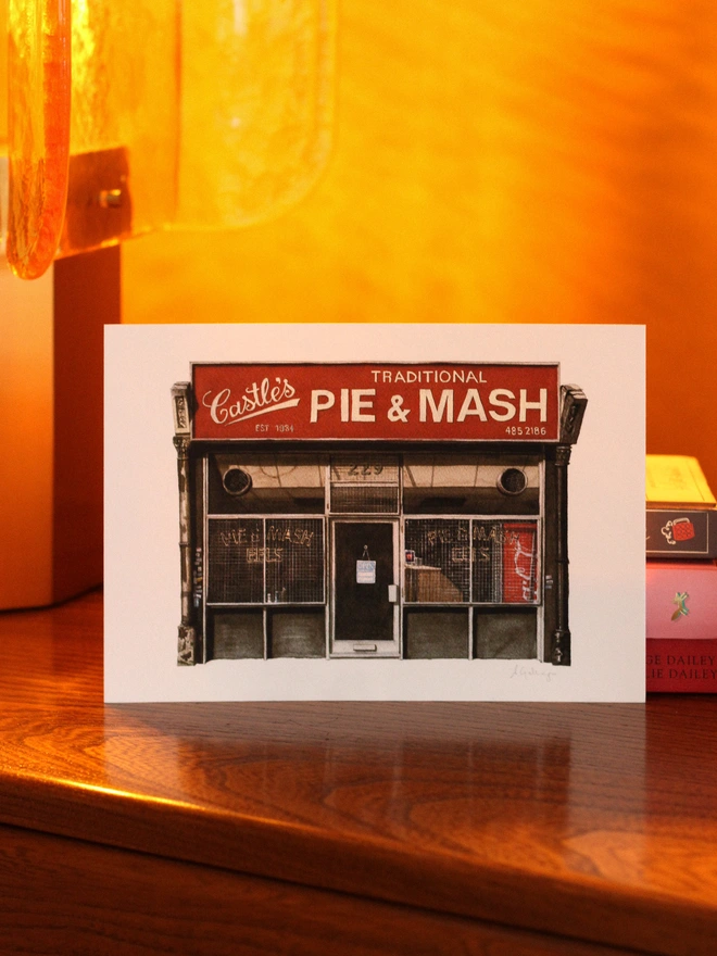 An architectural illustration of Castle's Traditional Pie & Mash shop front on a print, displayed on a wooden surface under warm amber lighting next to a stack of books.