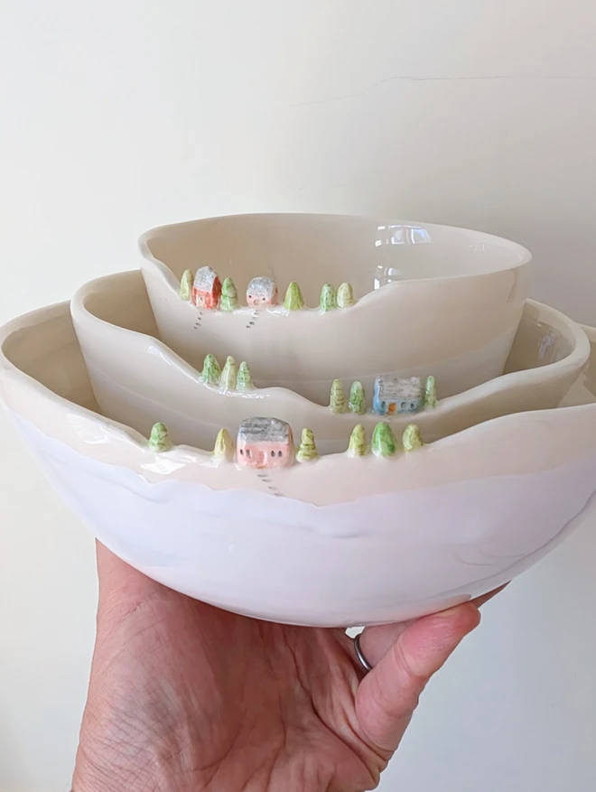 Handmade ceramic white nesting bowls with tiny house and tree details on the rim of all three bowls nesting inside each other held aloft by a hand