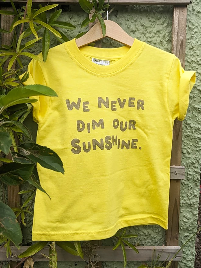 A yellow t-shirt with the slogan We Never Dim Our Sunshine hanging on a garden trelis