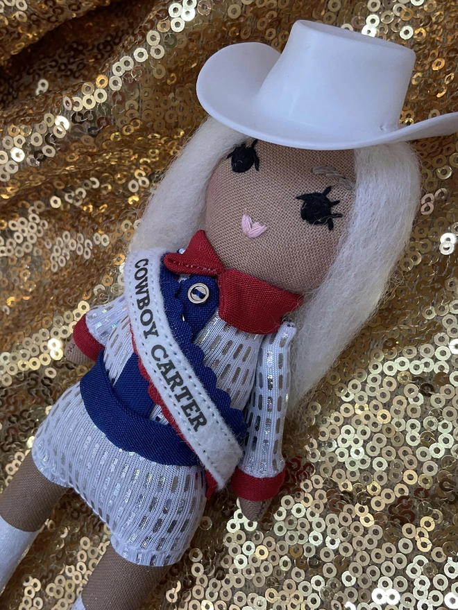 Small decorative fabric doll dressed like Beyoncé in her Cowboy Carter era. Red white and blue outfit.