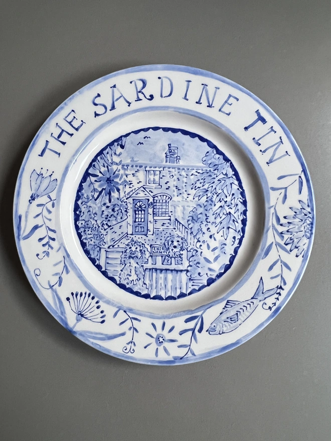 White plate illustrated in blue with "The Sardine Tin" on the rim with flowers and a fish and a house and 2 dogs inset
