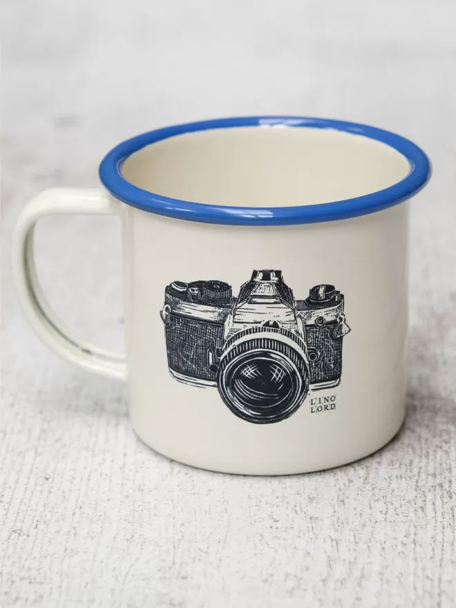 Picture of a Cream Enamel Mug with a Blue Rim with a Camera design etched onto it, taken from an original Lino Print